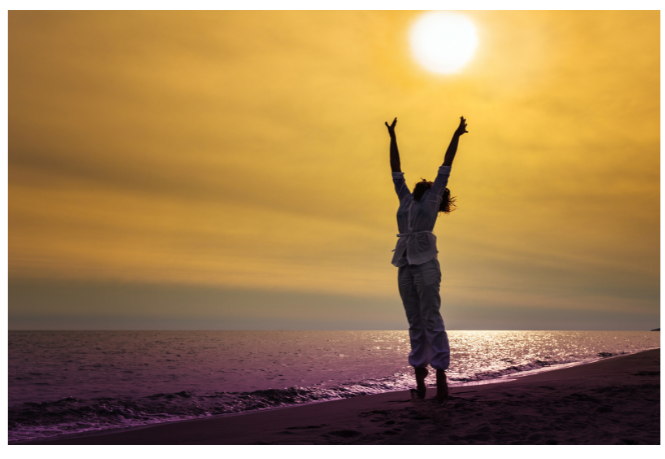 A woman jumping into the air with her hands raised during a sunset on the beach.