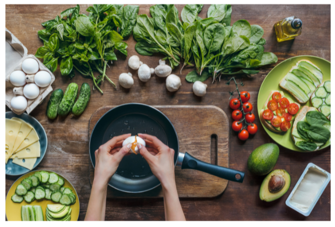 Top-down view of hands cracking an egg into a pan surrounded by vegetables and ingredients.