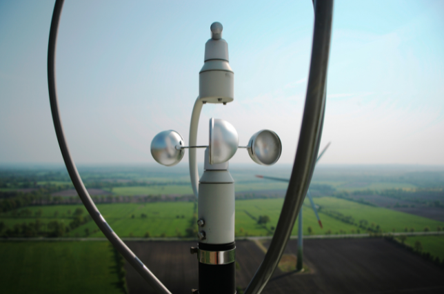 Close-up of a cup anemometer on a mast, with fields and a wind turbine blurred in the background.