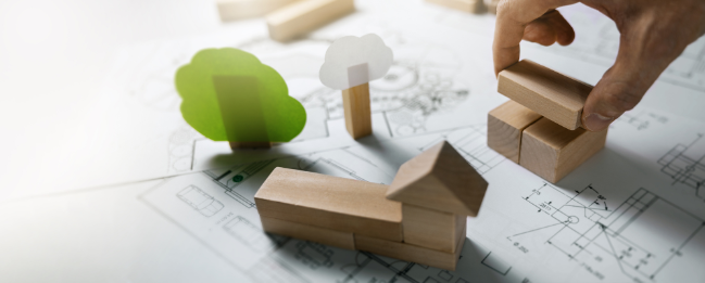 Hand arranges wooden blocks on an architectural plan with small tree cutouts—eco-friendly planning concept.