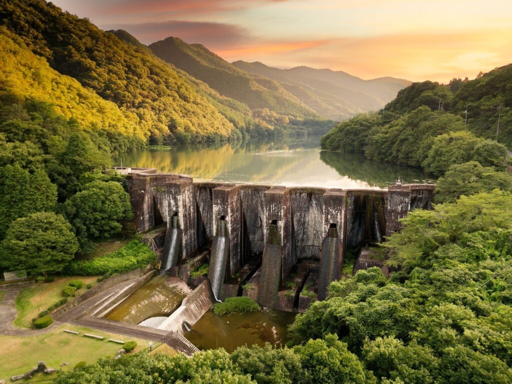 Stone dam amid lush green hills, holding a calm reservoir at sunset.