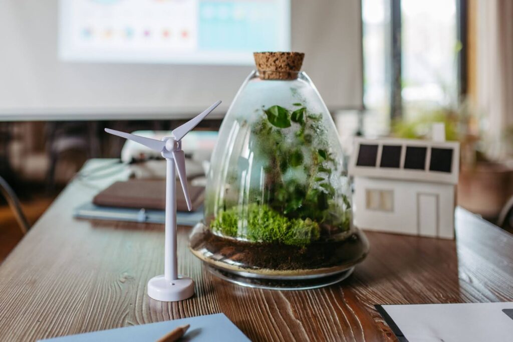 Mini wind turbine model beside a glass terrarium and a model house with solar panels on a desk.