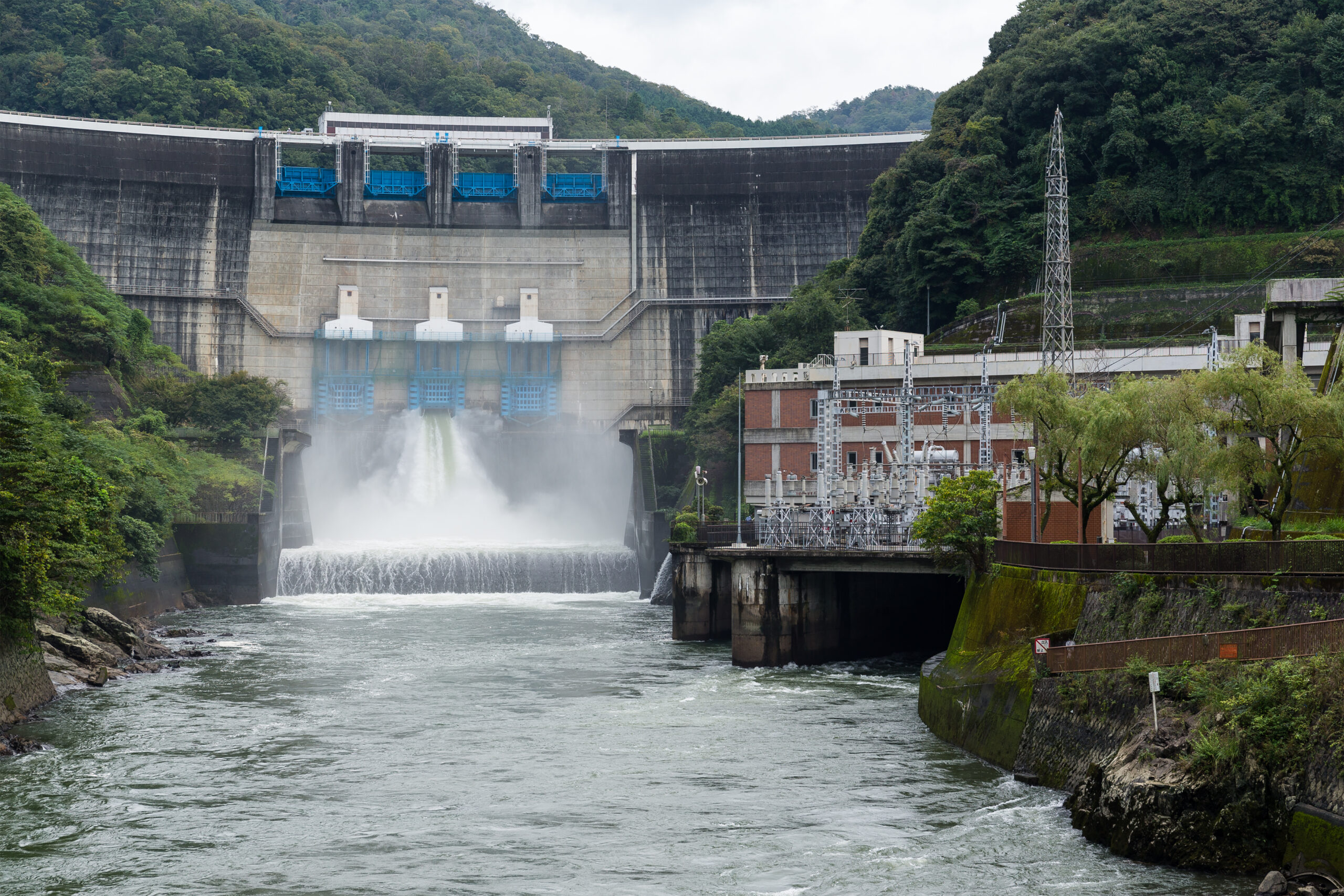 Hydroelectric dam releasing water into a river, with a nearby power station and forested hills.