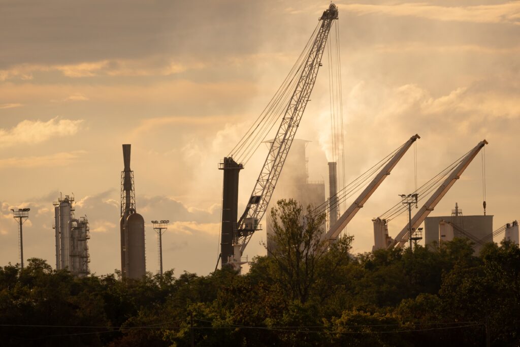 Industrial plant with tall stacks and cranes, steam rising behind trees under a hazy sky.