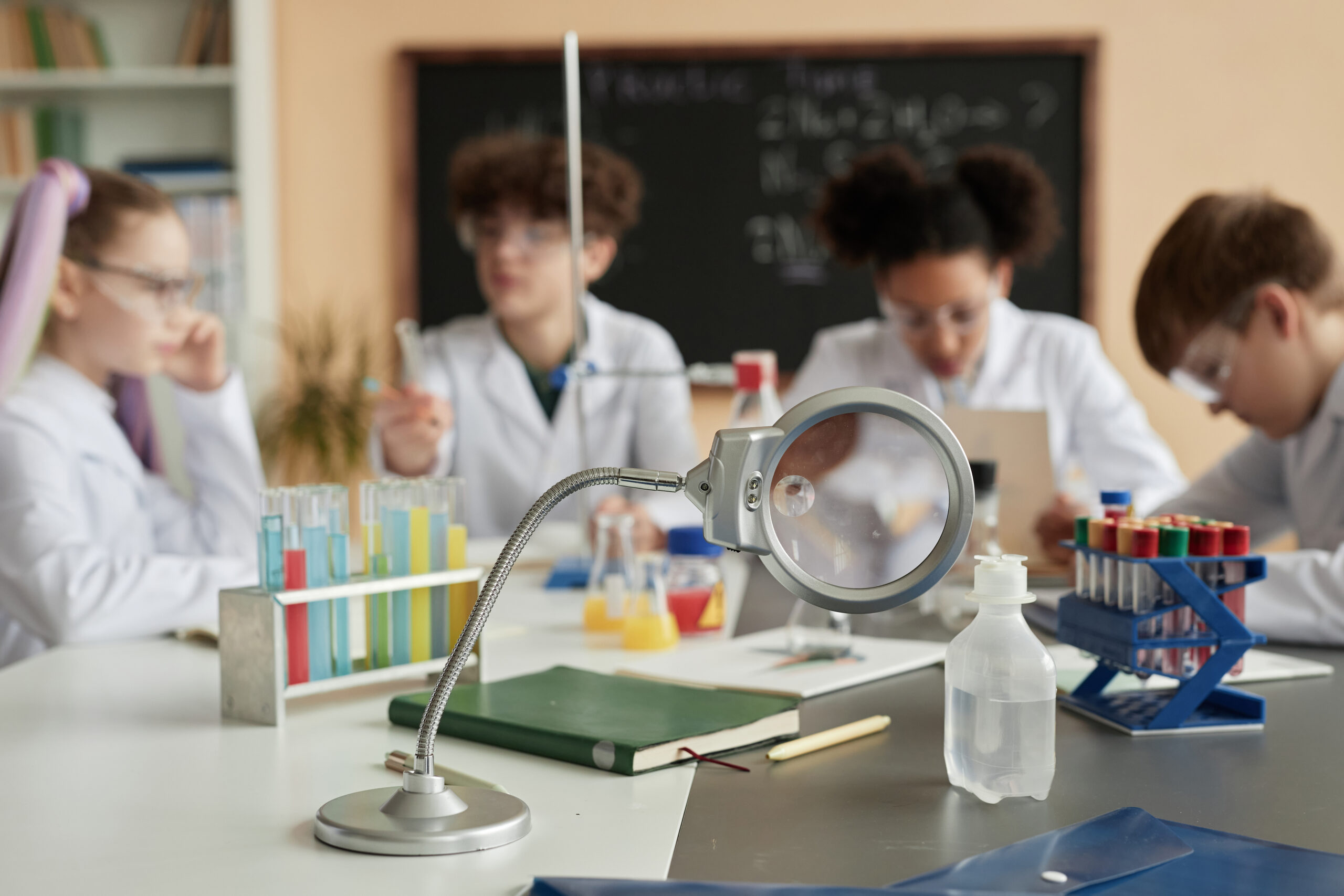 Students in lab coats and protective glasses with laboratory equipment on the desk.